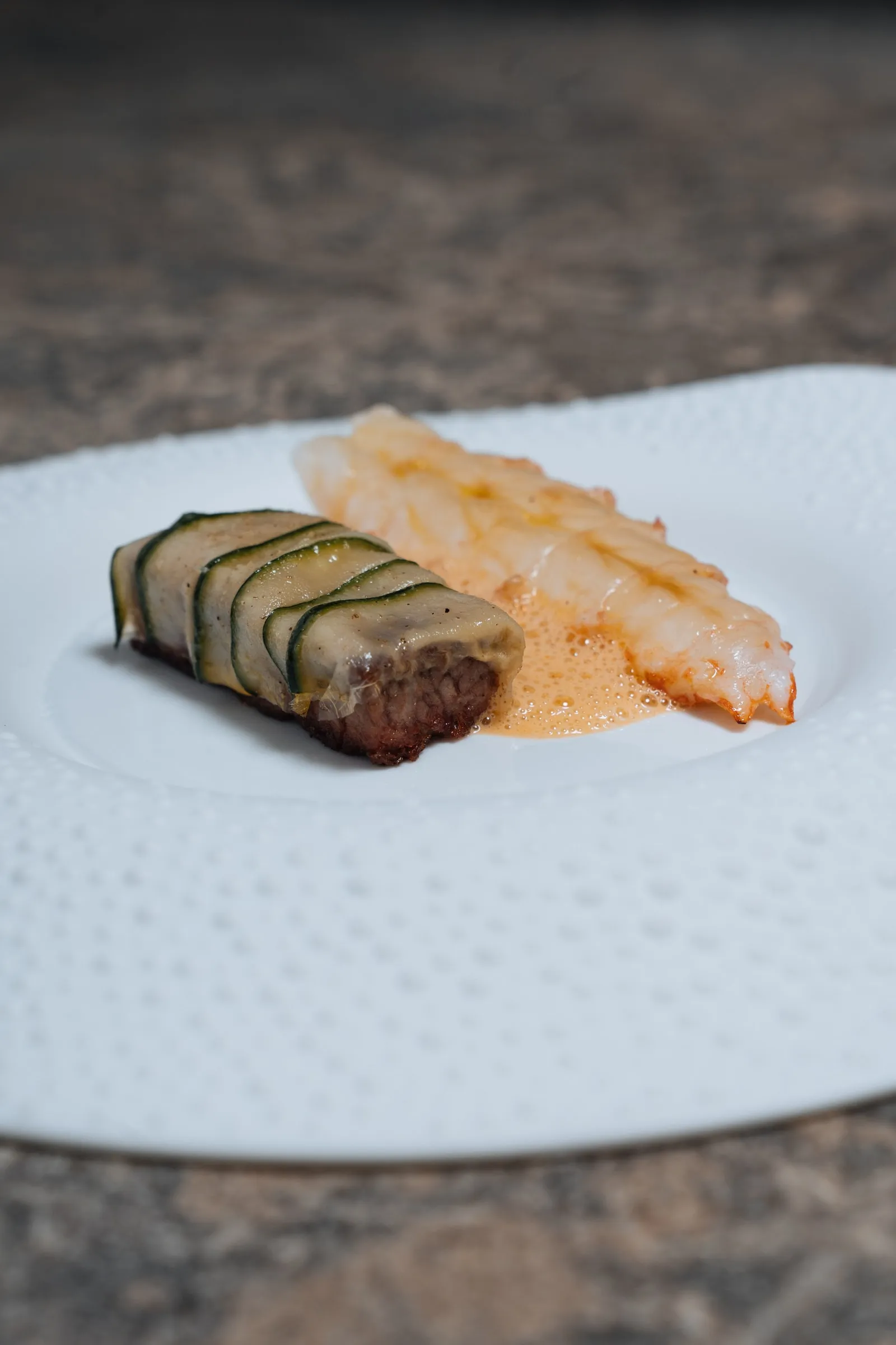 Nek van Tajiri gerold in courgette naast een langoustine, met saus op een wit bord